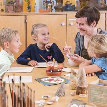 Tafelmalen Kinder ©Manfred Rauscher 2018 Eine Museumspädagogin mit vier Kindern an einem Tisch auf dem verschiedene Materialien zur Tafelmalerei liegen.
